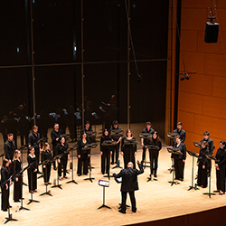 Two rows of singers stand in a semicircle, led by a conductor on a tall stage with floor to ceiling windows behind them.