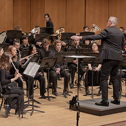 A band of woodwinds, brass, and percussionists play as a conductor leads.