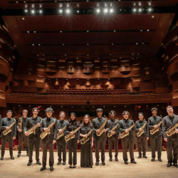 A large saxophone ensemble poses onstage with their saxophones.