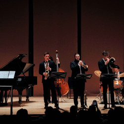 Three jazz musicians play onstage: a saxophonist, a trombonist, and a trumpeter. An upright bassist, drummer, and pianist play in the background.