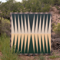 A painting with a symmetrical, geometric design of beige and dark green triangles is displayed in a naturally lush outdoor environment, surrounded by green plants, trees, and a pile of twigs.