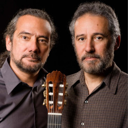 Sérgio and Odair Assad on a park bench with guitars.
