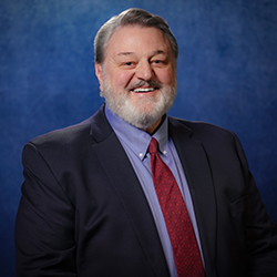Stanford Olsen standing against a blue background, wearing a suit and tie