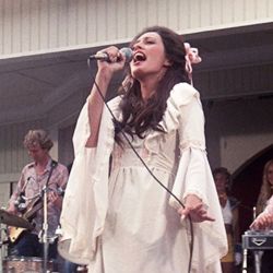 A woman with brown hair and light skin wearing a long white dress sings on stage